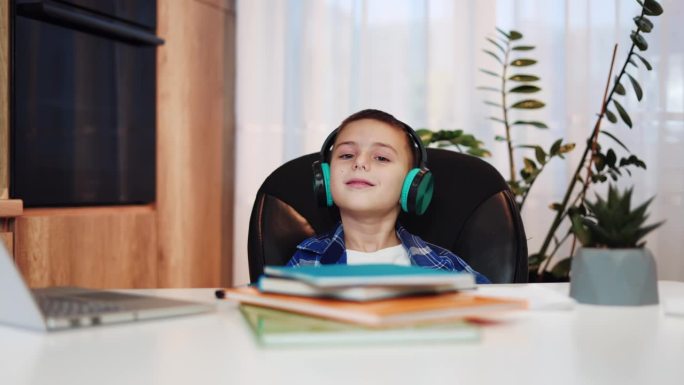 活跃的孩子戴着无线耳机听流行音乐，坐在课桌前摇着头打拍子。有趣的男孩从无聊的家庭作业中放松下来，独自
