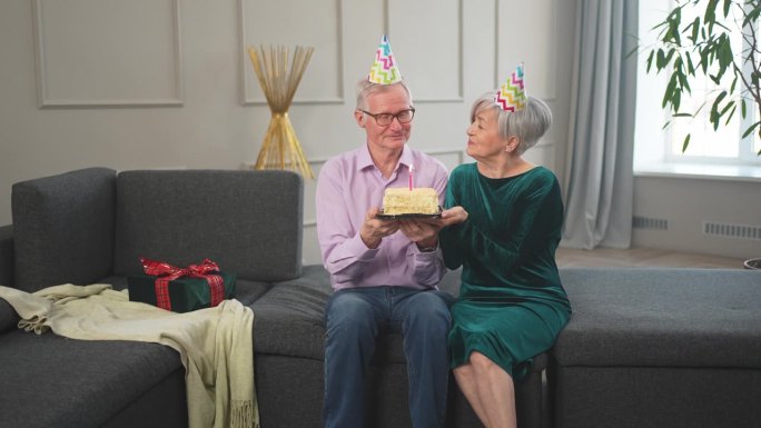 许个愿。家庭老年夫妇戴着派对帽在家里一起庆祝生日纪念日。老太太吹灭了生日蛋糕上燃烧的蜡烛。老人祝妻子