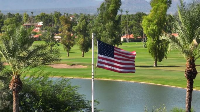 美国西南部棕榈树丛生的豪华乡村俱乐部高尔夫球场前飘扬的美国国旗。空中上升的照片显示湖泊和果岭。