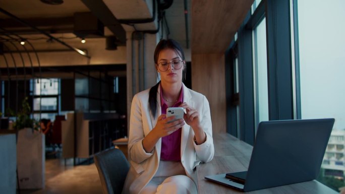 一个戴着圆眼镜的黑发女孩的肖像，穿着白色夹克和粉色衬衫，在一间现代办公室的全景窗旁的白色电话里工作
