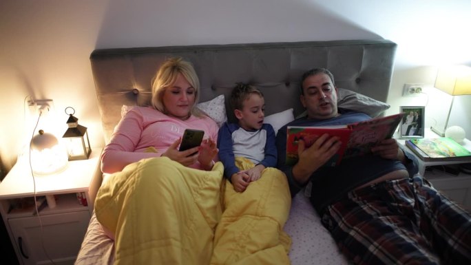 女人用手机定闹钟，而她的丈夫和儿子在床上看书