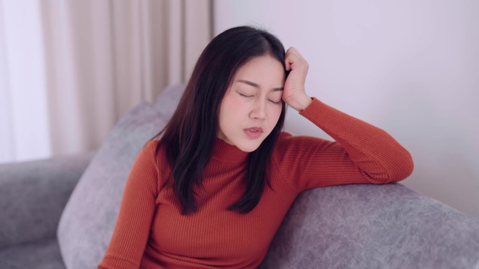 一名亚洲女性坐在家里的沙发上，因为焦虑而感到压力和头痛