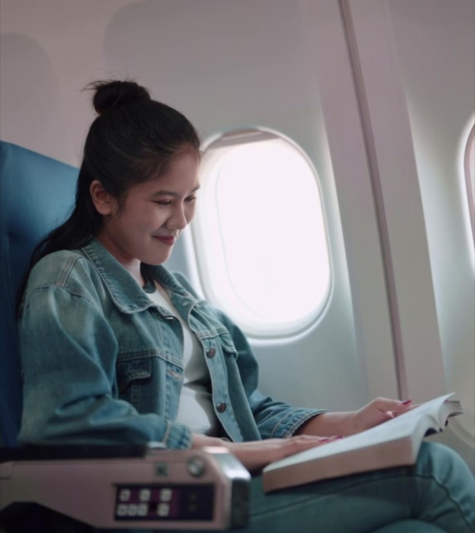 亚洲女性旅行者在飞往周末度假的飞机上享受她最喜欢的书。沉浸在她悠闲的旅程中，结合阅读的乐趣和长假的兴