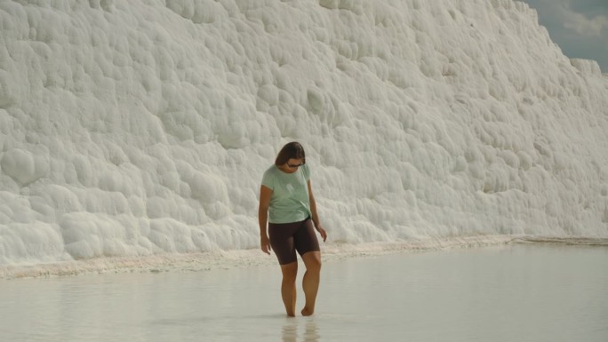 一位年轻女子在Pamukkale白色石灰华的泉水中浸泡她的脚。在背景中，有一堵白色的墙，上面有方解石