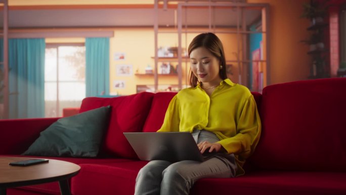 美丽的亚洲女人坐在舒适客厅的沙发上，在家里使用笔记本电脑。女性在社交媒体上微笑聊天、网购、看新闻