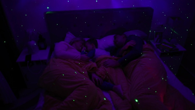 白人父母带着儿子睡在床上，床上有一盏白噪音RGB灯，灯上的星星在墙上投射
