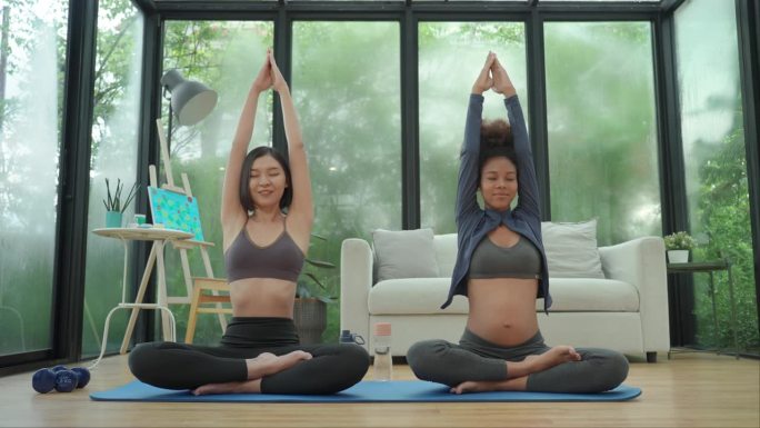 一位年轻的非洲孕妇在亚洲瑜伽教练的帮助下，在家中进行瑜伽练习，教授缓慢而感恩的运动，以改善健康的生活