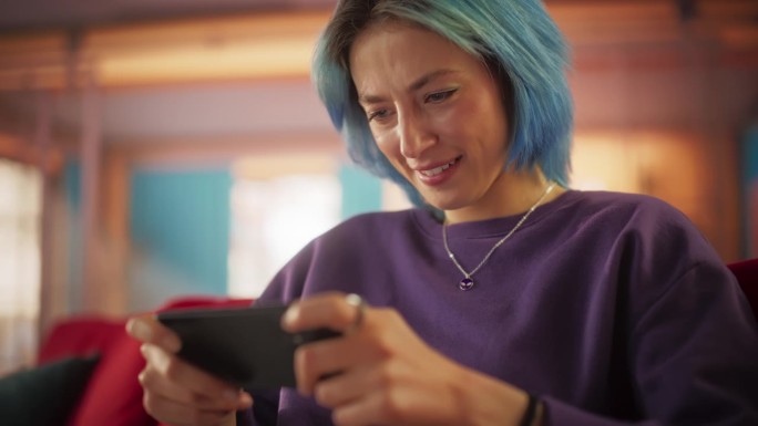 美丽时尚的蓝头发女人在她色彩缤纷的公寓里用智能手机玩电子游戏的慢动作肖像。女性青少年在在线合作游戏中