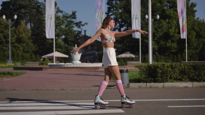 在一个阳光明媚的日子里，一个戴着眼镜的年轻女子穿着溜冰鞋，在城市公园的背景下优雅地跳舞。缓慢的运动。