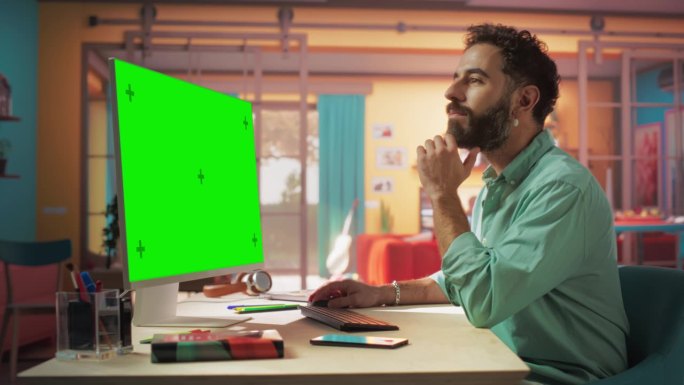 年轻英俊的男子从家里工作的桌面电脑与绿屏模拟显示。创意男查看社交媒体，浏览互联网。明亮阁楼公寓的客厅