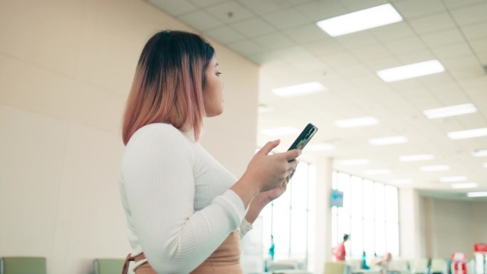 自信的年轻亚洲短头发女子使用智能手机，查看航班信息板，坐在国际机场，快乐的女乘客带着旅行行李等待旅行