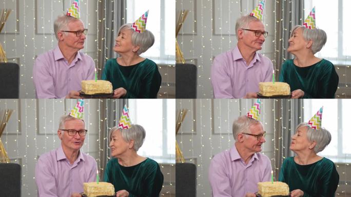 许个愿。家庭高级夫妇戴着派对帽在家里一起庆祝生日纪念日。老太太吹灭了生日蛋糕上燃烧的蜡烛。老人祝妻子