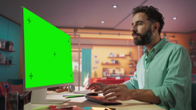 年轻英俊的男子从家里工作的桌面电脑与绿屏模拟显示。微笑的男性查看公司账户，给同事发信息，做创意项目