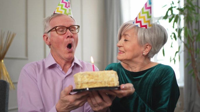 许个愿。家庭老年夫妇戴着派对帽在家里一起庆祝生日纪念日。老人吹灭了生日蛋糕上燃烧的蜡烛。老太太祝丈夫