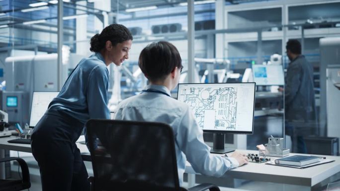 西班牙裔女经理和亚裔男工程师在拥有机械臂的自动化工厂使用带有微芯片蓝图的台式电脑。同事们讨论电子产品