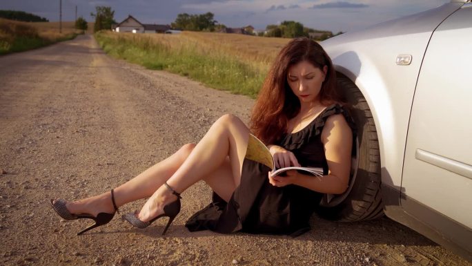 穿着黑色晚礼服的美女在肮脏的乡间小路上倚着车轮看书