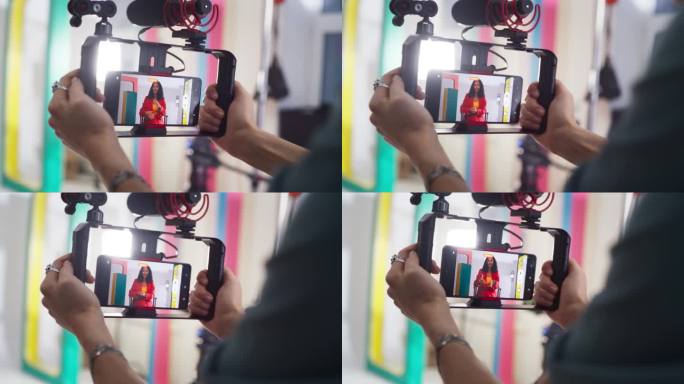 在智能手机上拍摄一位穿着彩色衣服的女电视主持人:时尚的内容创作者与摄影师一起为她的在线频道制作视频的