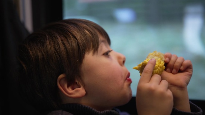 小男孩在火车上吃玉米当零食，乘客小孩一边吃东西一边盯着路过的风景