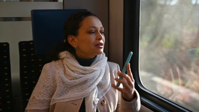 坐火车旅行的年轻女子，一边用智能手机聊天，一边梦幻般地望着窗外。坐火车的女乘客。人。交通工具。旅行