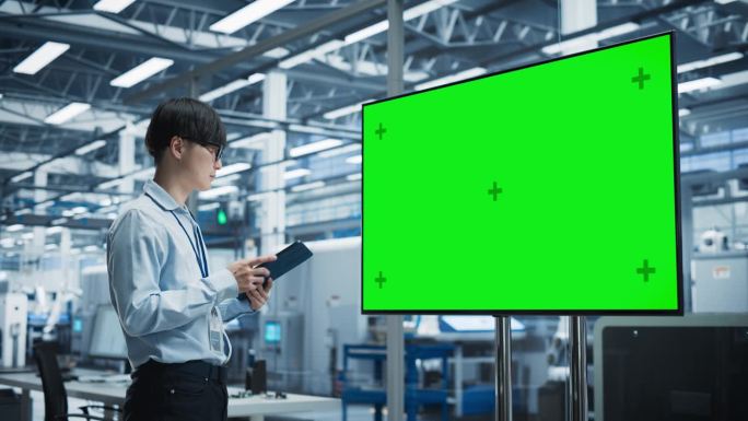 在拥有机械臂的自主电子厂，亚洲男性首席工程师一边使用平板电脑，一边看绿屏Chromakey电视。一个