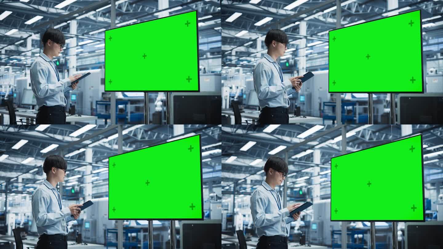 在拥有机械臂的自主电子厂，亚洲男性首席工程师一边使用平板电脑，一边看绿屏Chromakey电视。一个