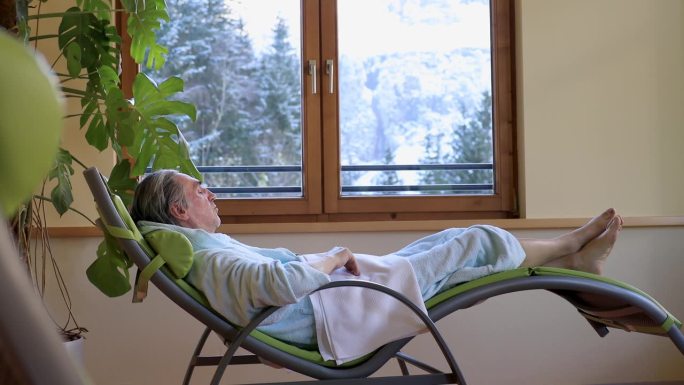 疗养院里的一间休闲室，一个头发花白、穿着浴袍的男人躺在椅子上