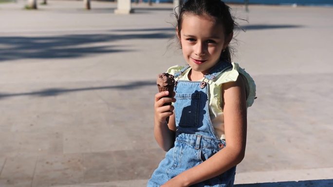 漂亮的小女孩穿着牛仔背心裙，在户外吃着美味的巧克力冰淇淋
