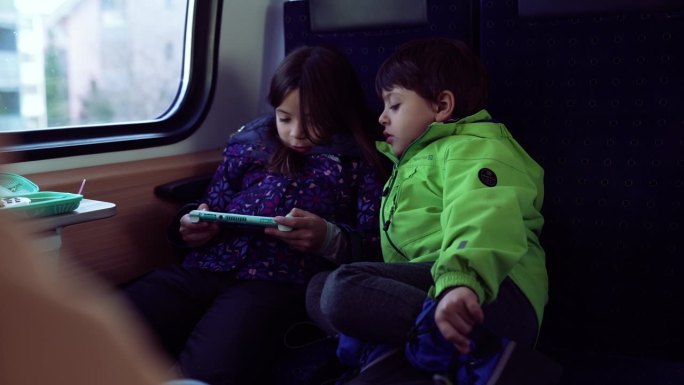 乘客小女孩拿着掌上游戏机玩游戏，弟弟盯着她的屏幕玩，孩子们在火车里玩科技