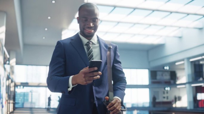 穿着完美剪裁西装的年轻黑人男子的肖像走在公司办公室的走廊上，他微笑着使用智能手机。成功的商人通过短信