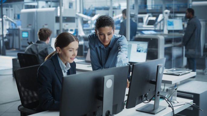 白人女工程师和西班牙裔产品经理使用台式电脑，在自动化工厂用机器手说话。不同的同事讨论新的高科技设备生
