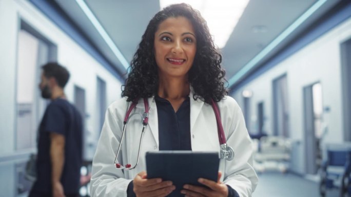 一位态度积极的女医生，平静地走过医院走廊，使用数码平板电脑，向护士和同事打招呼的跟踪镜头。明亮现代新