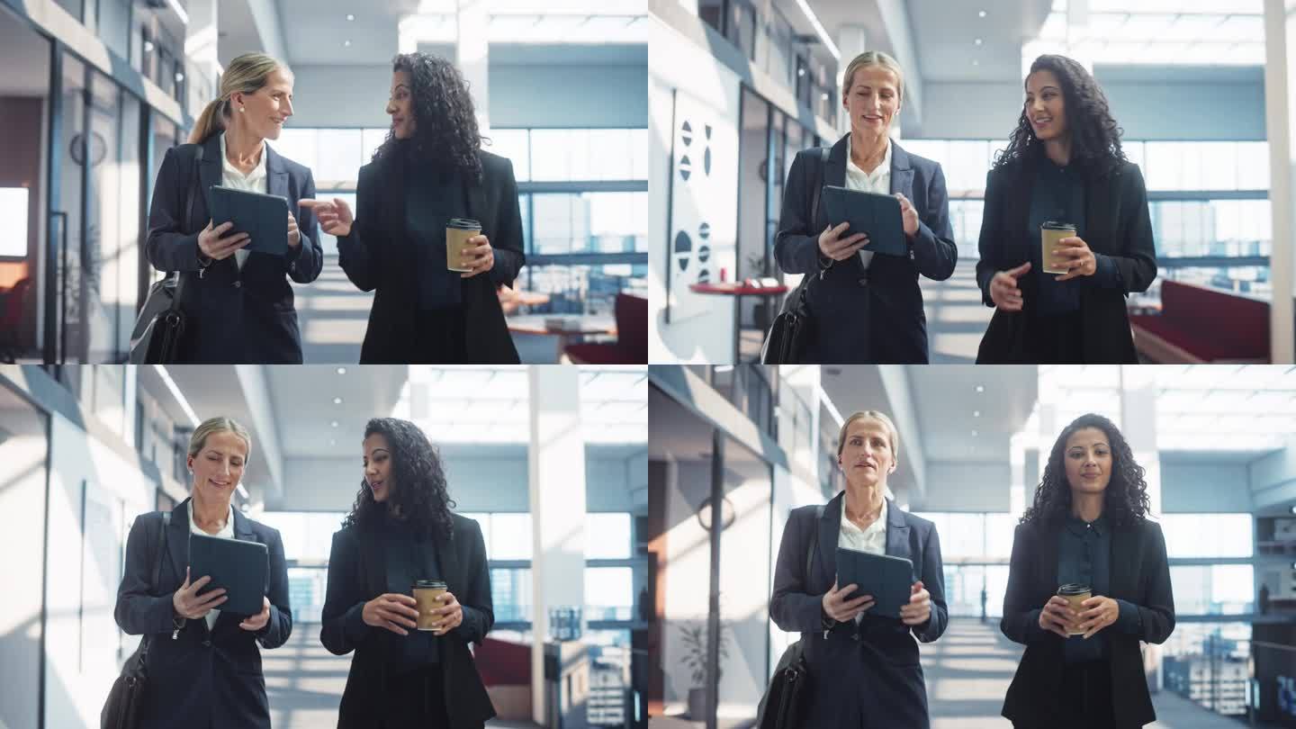 优雅的女商人走在明亮的办公大楼里，她们用平板电脑讨论业务。聪明自信的女人在上班的路上开个简短的会