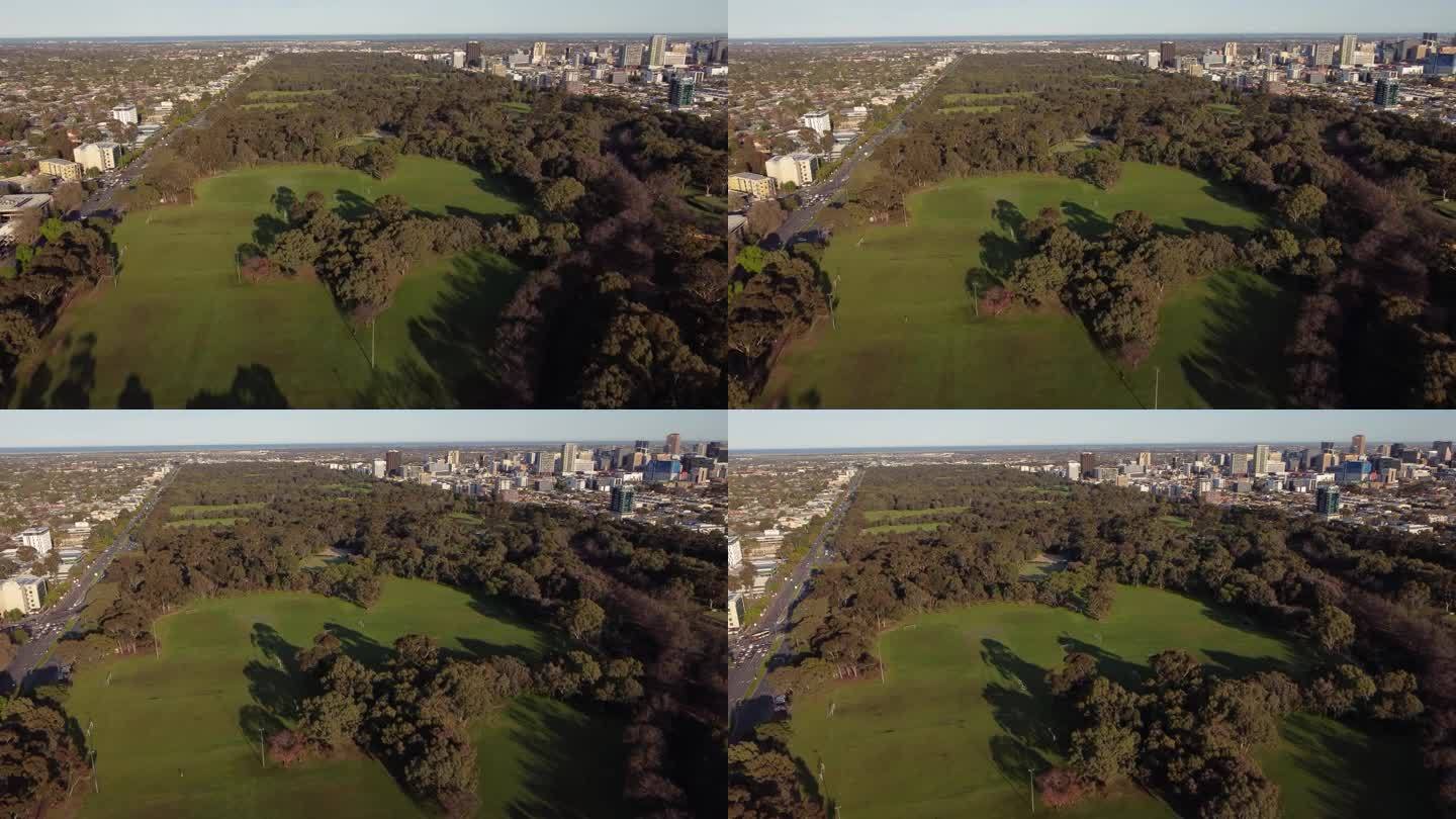 一架飞机飞过阿德莱德公园，转向阿德莱德市。绿色椭圆和树木可见，背景是城市建筑。