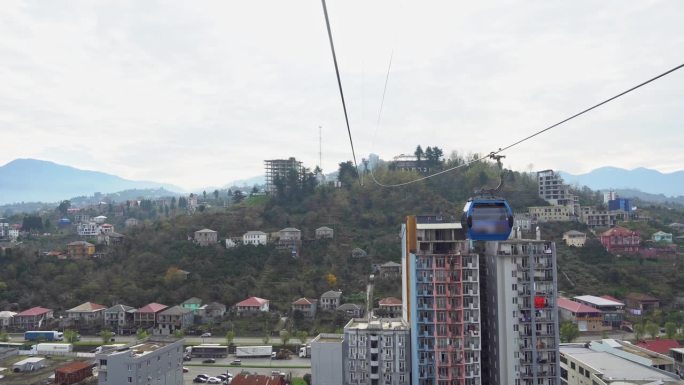 从索道的船舱里，沿着缆索上升，俯瞰城市，背景是群山。悬挂架沿滚子接头移动。山地缆车