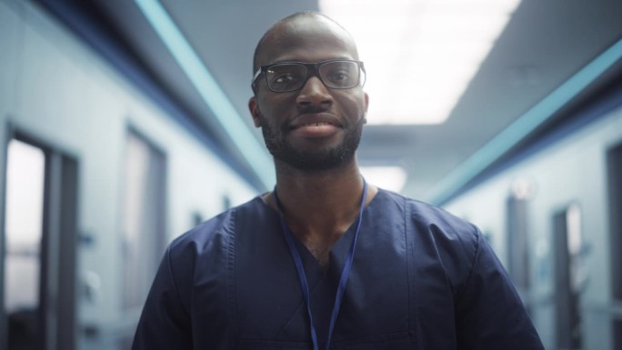 一名黑人医疗保健专业人员在诊所走廊摆姿势，微笑着看着镜头。戴眼镜的医生在医院工作，拯救生命