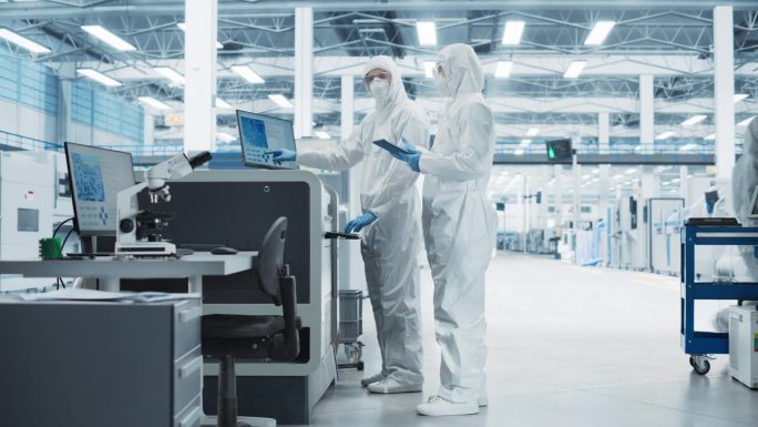 两位穿着无菌服的专家走在电子厂研究中心。手持平板电脑的科学家和接近焊锡喷墨打印机的工程师开始了电路板