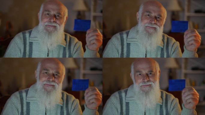 特写:一个穿着毛衣、留着胡子的老人，手里拿着一张蓝色卡片，咧嘴笑着。