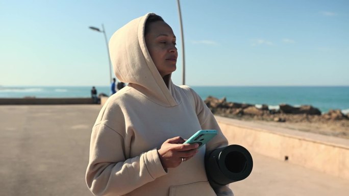 微笑幸福的女人站在大西洋海岸线上，使用智能手机，查看移动应用程序