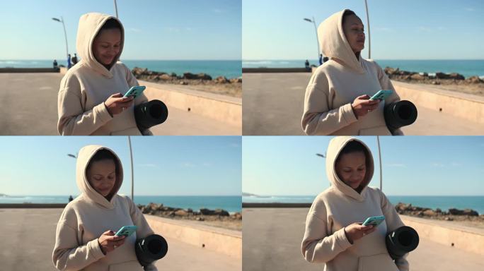微笑幸福的女人站在大西洋海岸线上，使用智能手机，查看移动应用程序