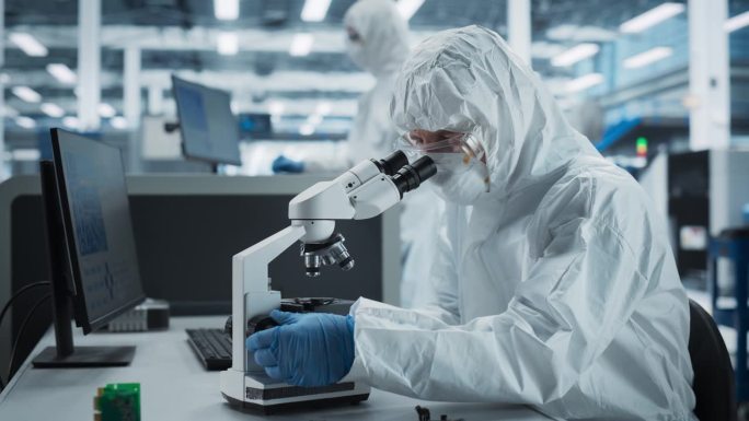 两位科学家在电子工厂的无菌套房:工程师使用显微镜和台式电脑检查微芯片，而他的同事使用焊接喷墨打印机打