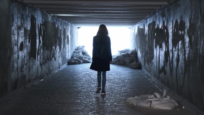 一个女人的剪影沿着一条变成防空洞的地下通道走向灯光。后视图，破旧的隧道，沙袋。乌克兰战争。
