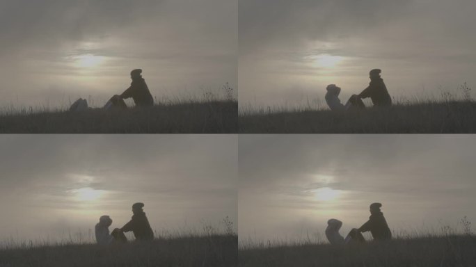 一名女子扶着一名男子的膝盖，在日落时被薄雾笼罩的山脉背景下，在干燥的秋草上练习按压肌肉。与自然融为一