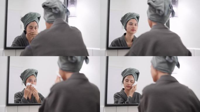 一位亚洲女性在洗面后，在浴室镜子前用毛巾轻轻擦干脸部。捕捉这个护肤程序的清爽形象