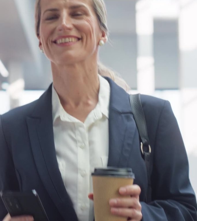 垂直屏幕:一个女人在使用她的智能手机，微笑着走在空荡荡的办公大厅里。一位穿着时髦休闲装的女商人，端着