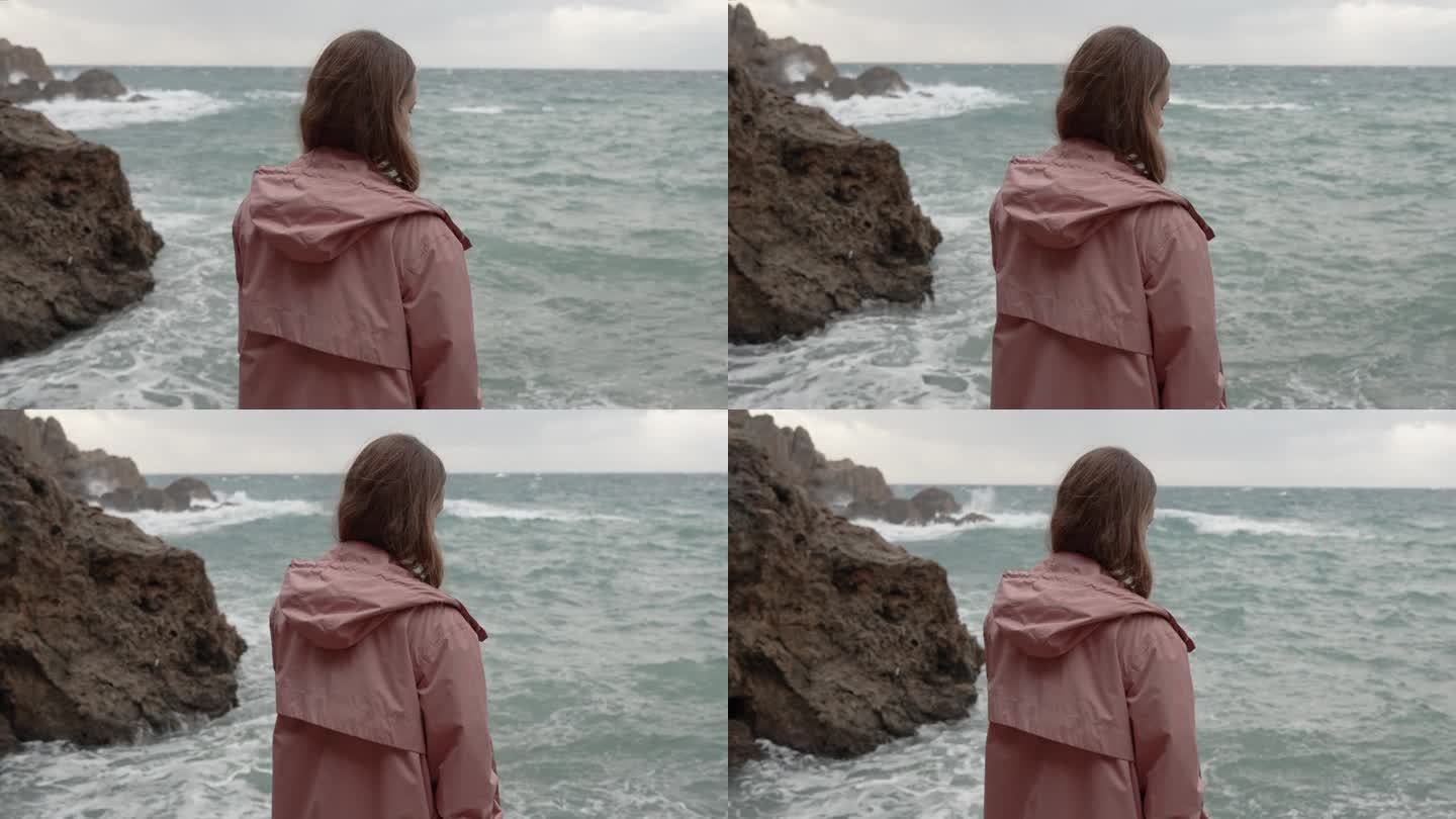 从背后看，一个年轻女子独自站在岩石上，面对汹涌的大海。