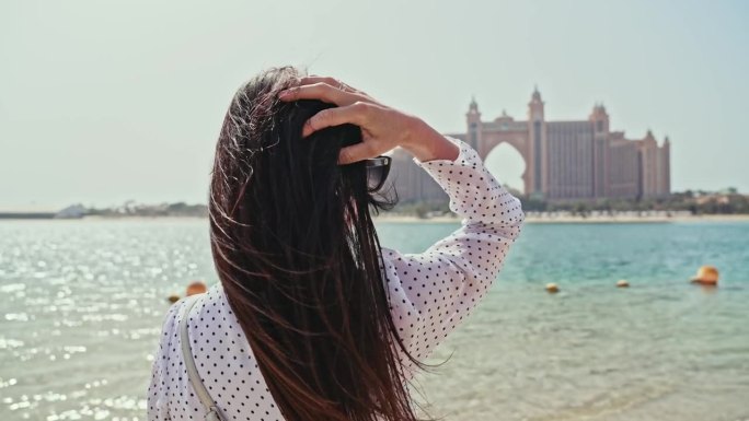 在迪拜五星级酒店的映衬下，女孩的头发在风中美丽地绽放。幸福的女人在城市的海边散步。度假和观光。阿联酋