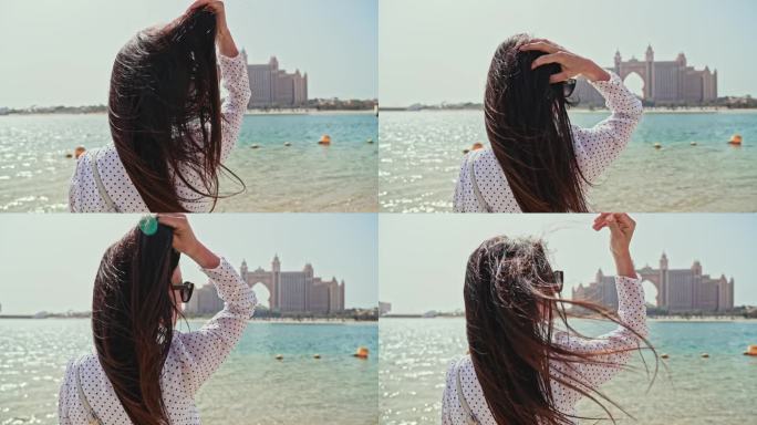 在迪拜五星级酒店的映衬下，女孩的头发在风中美丽地绽放。幸福的女人在城市的海边散步。度假和观光。阿联酋
