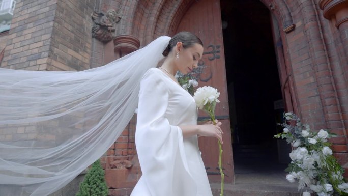 新娘身穿白色长裙，头纱手持白花。行动。年轻的女士站在敞开的教堂门口