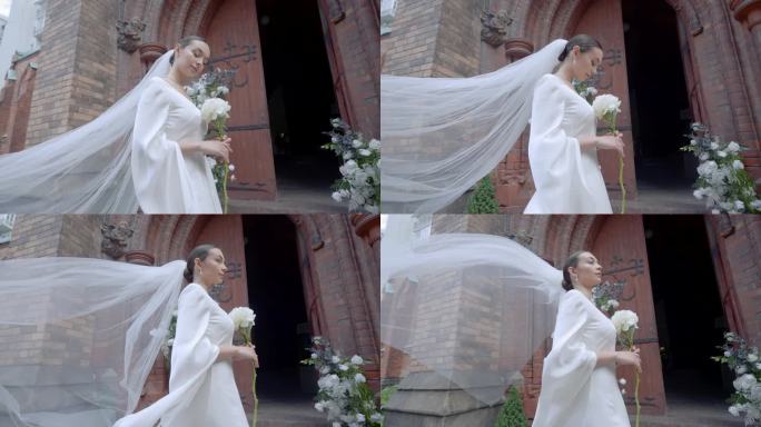 新娘身穿白色长裙，头纱手持白花。行动。年轻的女士站在敞开的教堂门口