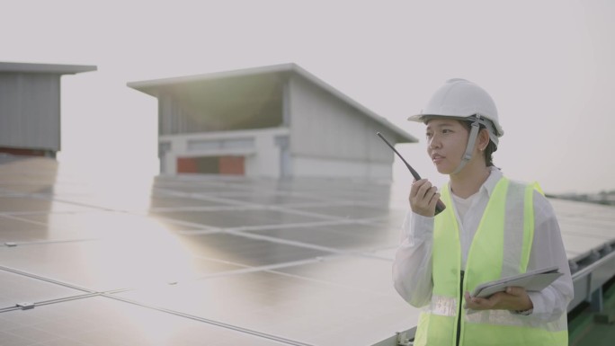 亚洲中年女工程师在太阳能农场使用数字平板电脑，穿着防护服，戴着头盔，通过无线电与她的团队沟通。